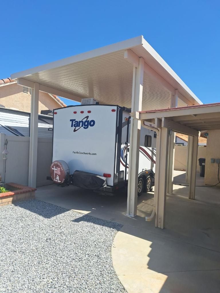 A camper trailer parked under a white carport on a gravel driveway, with a clear blue sky.