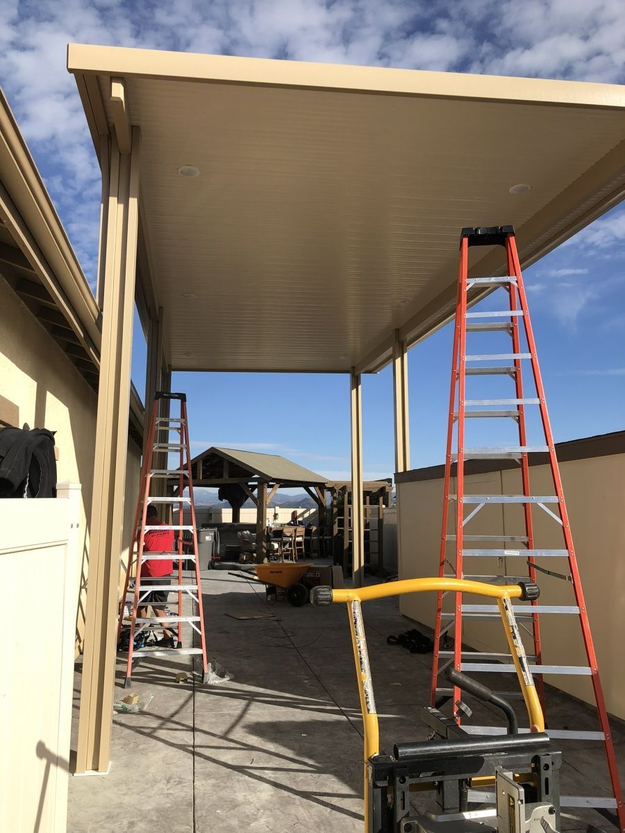 A tan-colored patio cover under construction. Two ladders are propped up. Construction materials lie on the ground.
