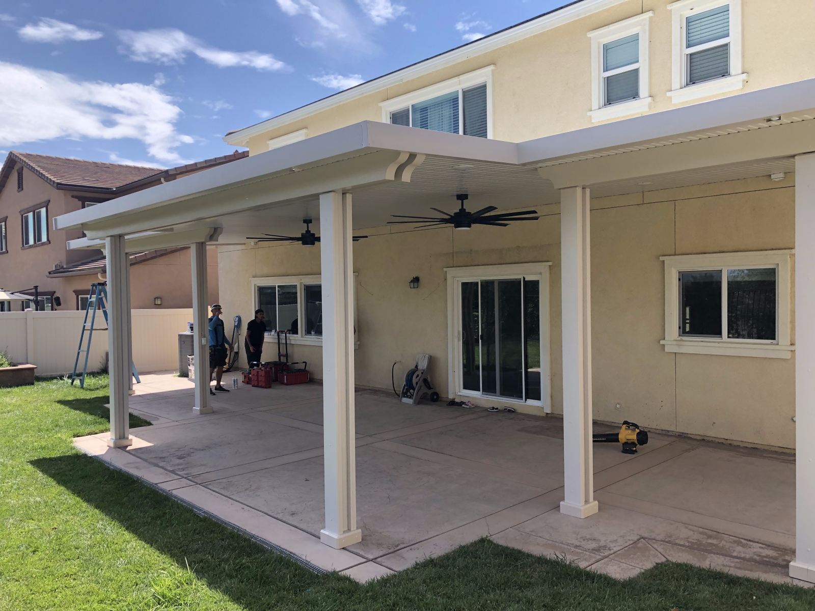 A covered patio extension of a house. Two people work, concrete patio, green lawn, blue sky.