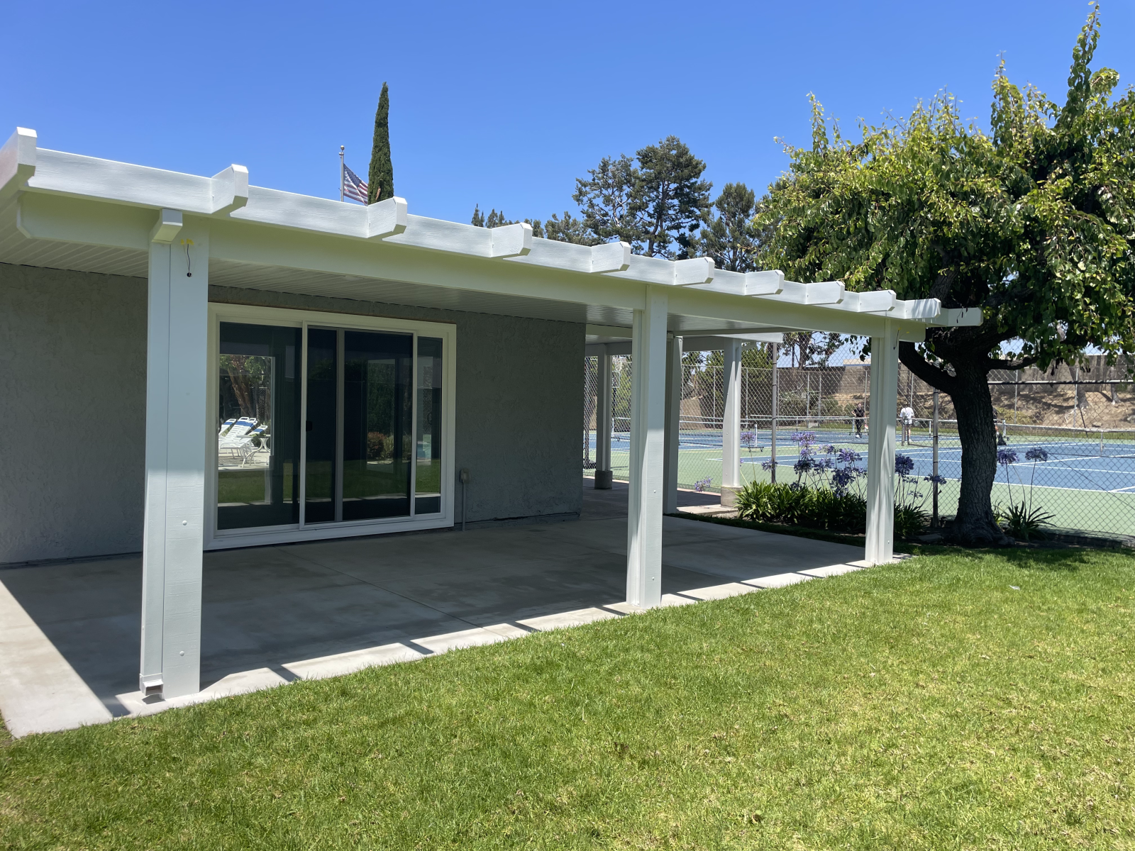 A white pergola covers a concrete patio next to a building with sliding doors. Green grass and a tennis court are nearby.