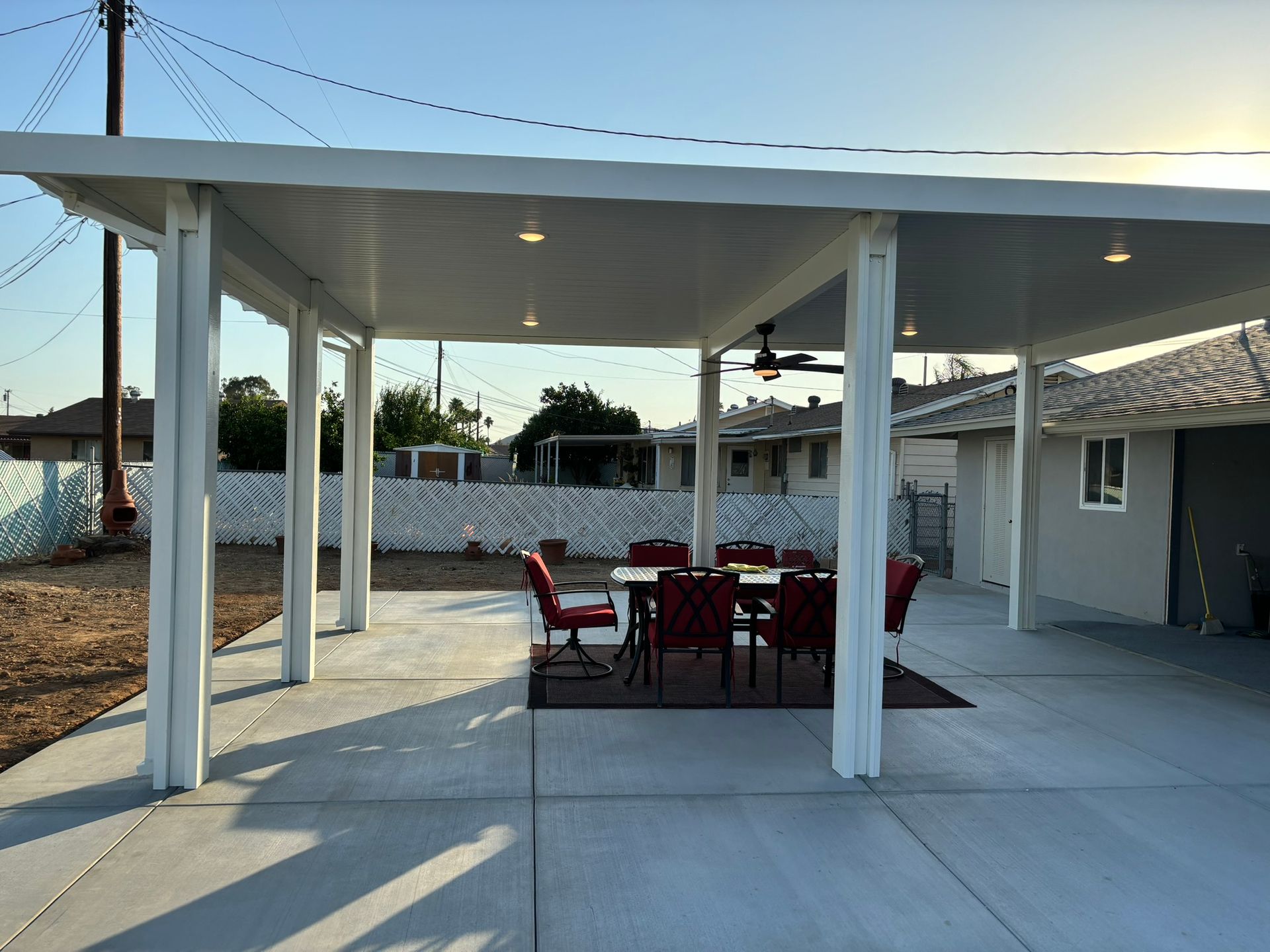 A white patio cover with recessed lights over a dining table with red chairs, on a concrete patio.