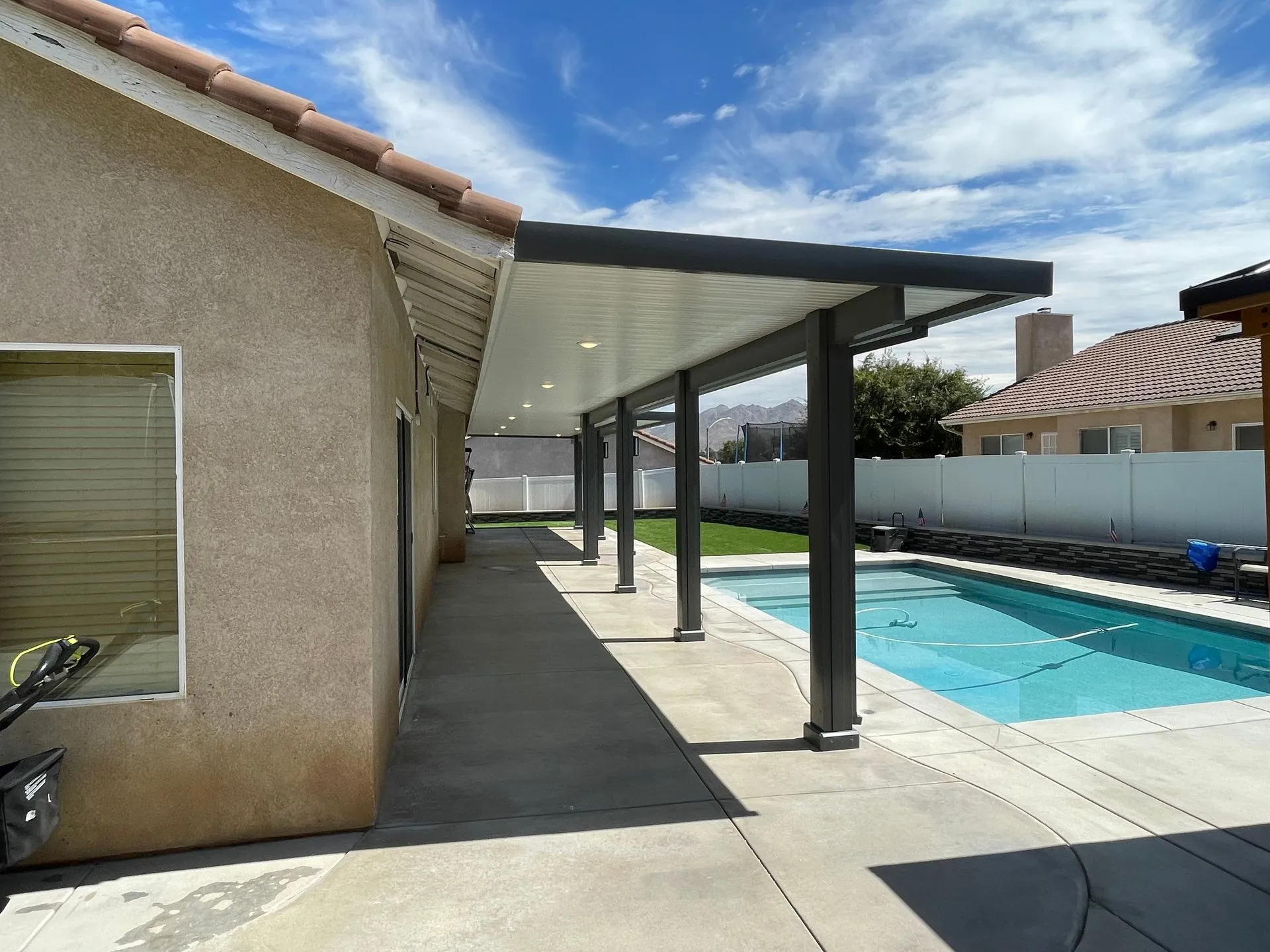 Patio cover extends from beige house.  Pool and grass visible.  Blue sky and white clouds.