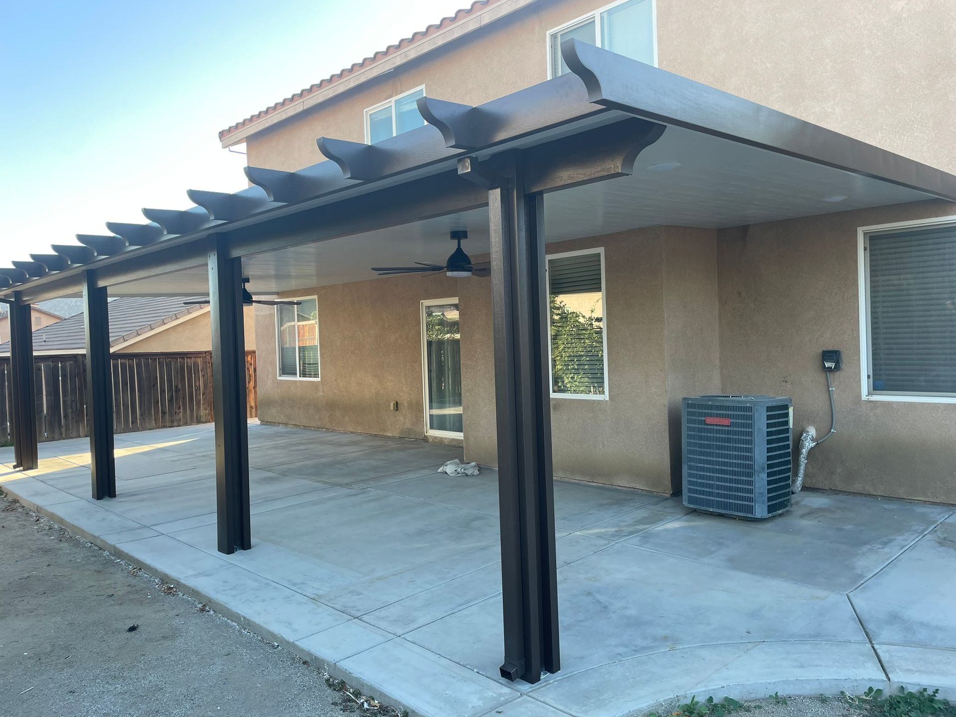 Brown patio cover attached to a beige house, with concrete flooring and dark support beams.