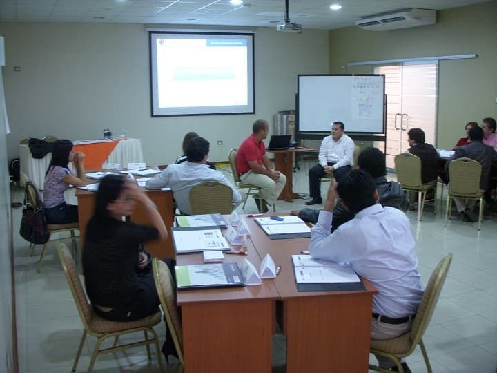 Un grupo de personas están sentadas en mesas frente a una pantalla de proyector.