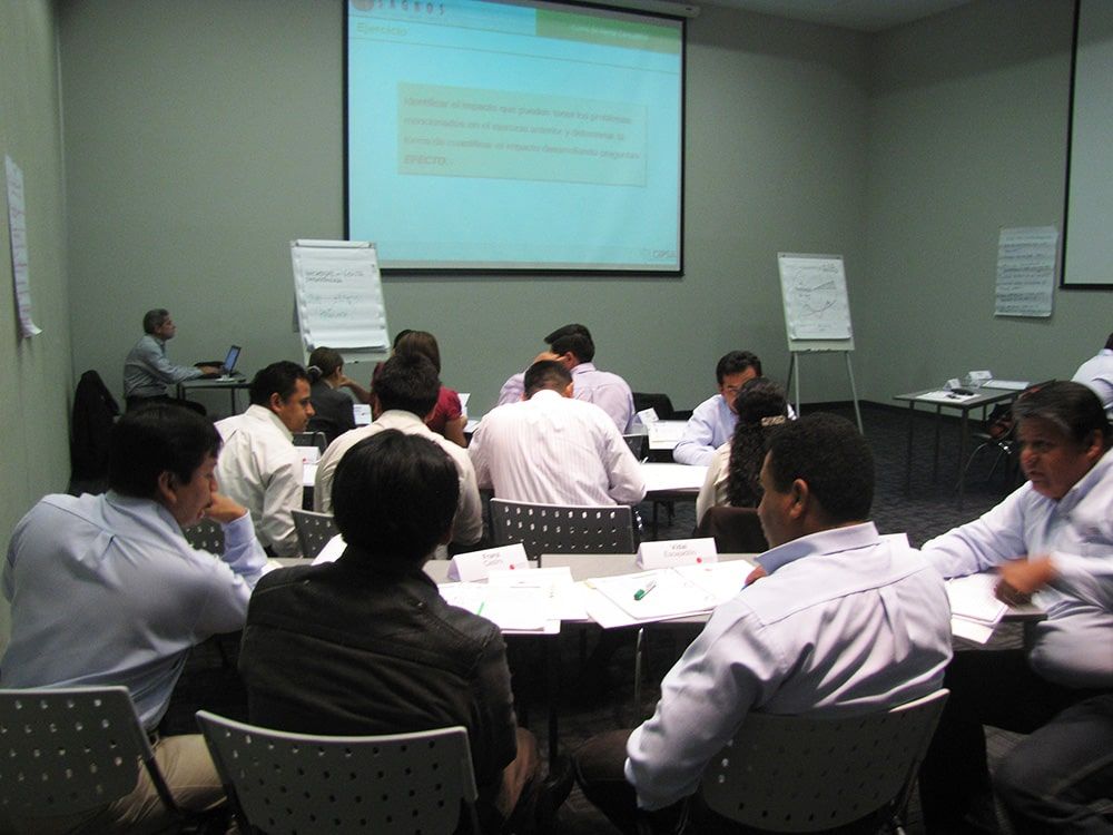 Un grupo de personas están sentadas en mesas frente a una pantalla de proyector.