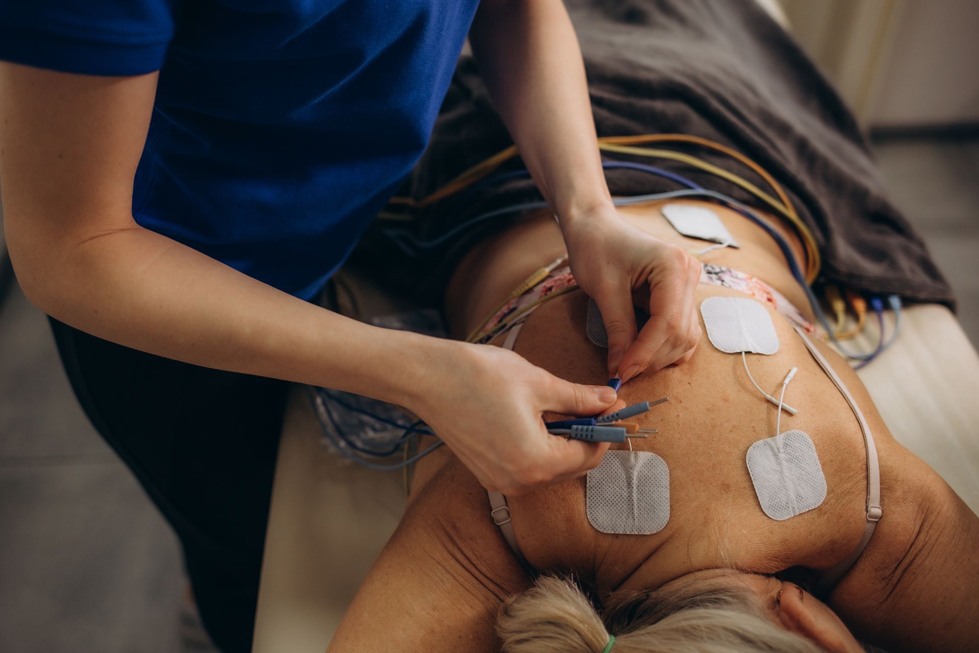 A practitioner in a blue shirt applies electrodes to a patient's back during a physical therapy treatment.