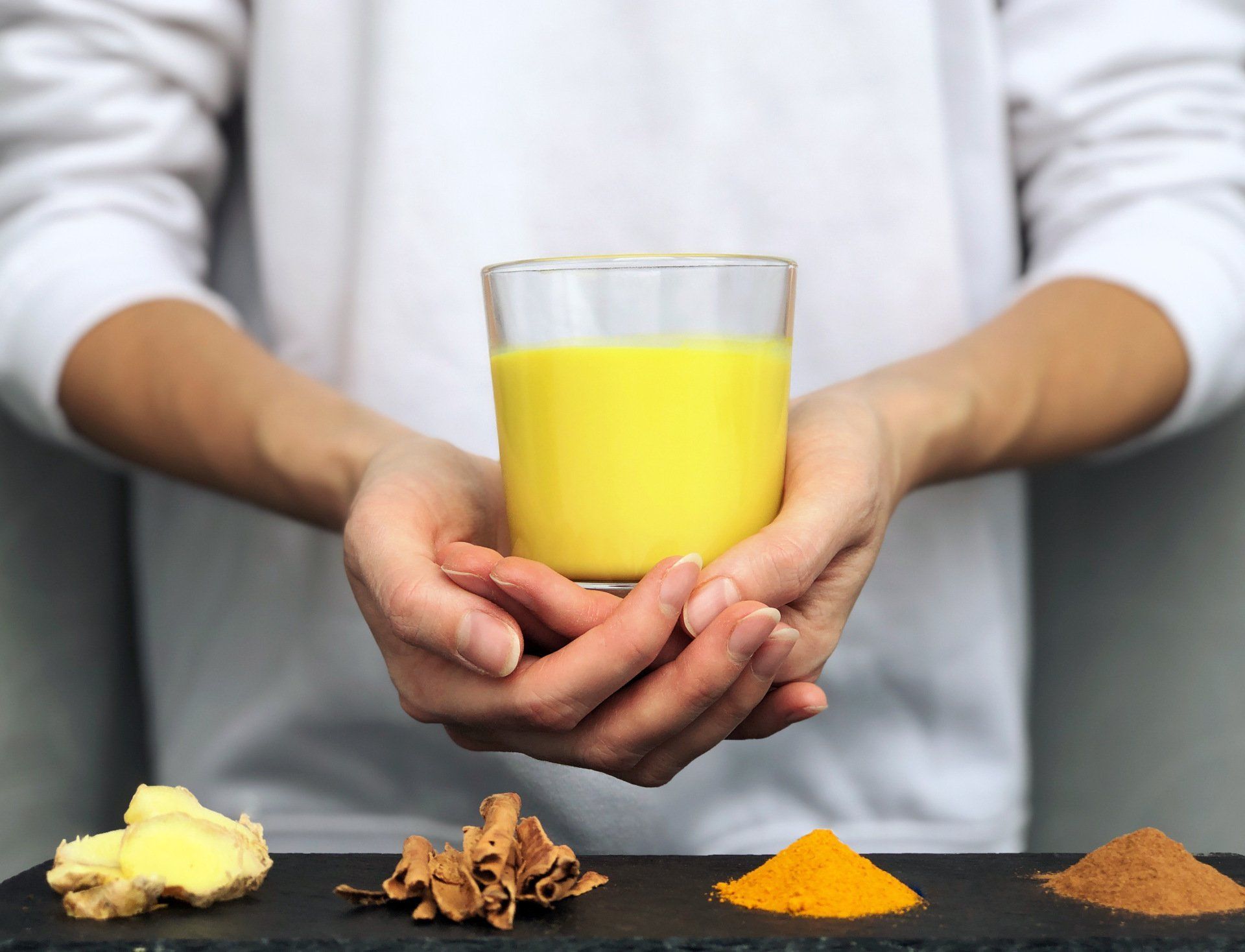 A person in a white long-sleeved shirt holds a glass of golden yellow drink behind piles of ginger, cinnamon, and turmeric.