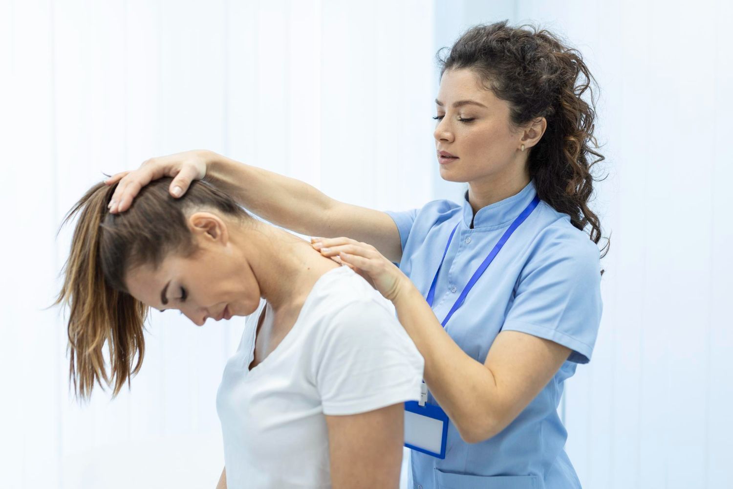 Woman in white shirt having neck examined by a healthcare professional in blue scrubs.