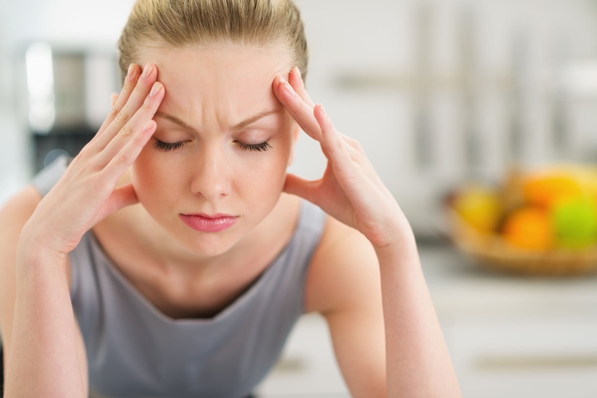 Woman with hands on temples, eyes closed, appearing stressed in a kitchen.