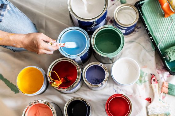 Hands dipping a brush into a blue paint can among open cans of colorful paint