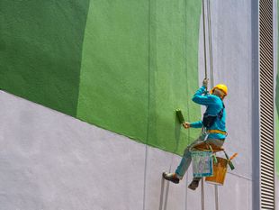 Worker painting a green and gray building wall while suspended by rope and bucket