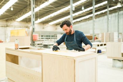 Woodworker sanding a light wooden cabinet in a bright workshop