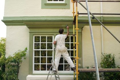 Painter on a ladder painting the exterior trim of a house under scaffolding.