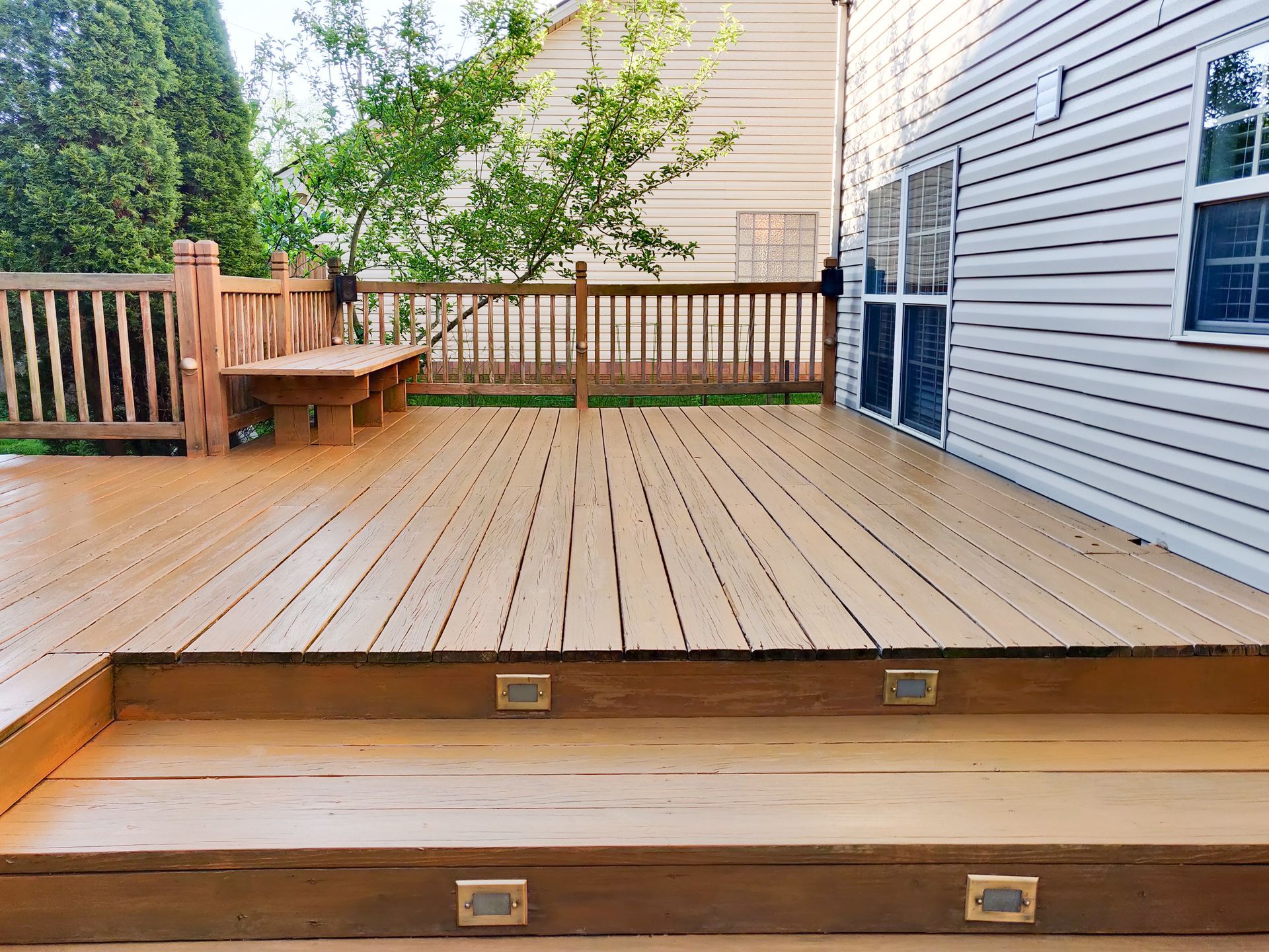 Wooden backyard deck with steps, railing, a bench, and a tree beside a house