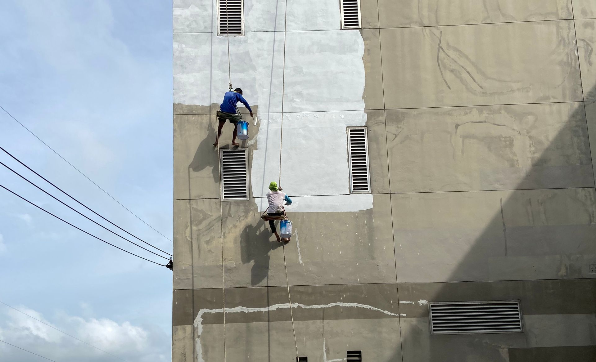Two workers on rope access inspect a tall building façade with windows and vents.