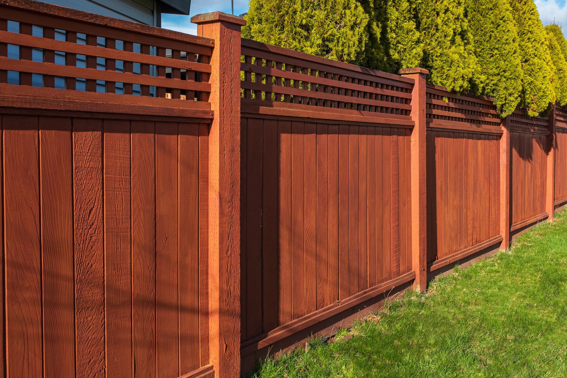 Red wooden privacy fence along a grassy yard, with lattice top panels and sunlight casting shadows.