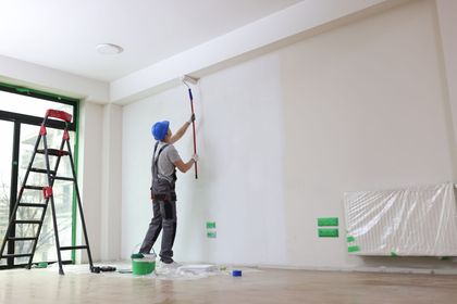 Worker painting a white wall inside a bright room, with ladder and paint supplies nearby