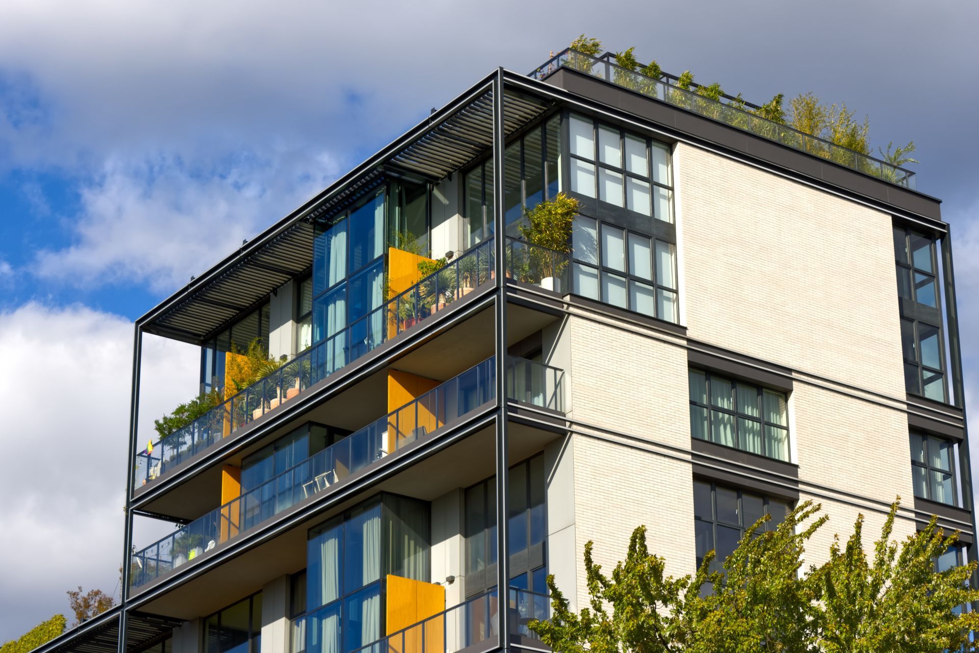 Modern apartment building with glass balconies, yellow accents, and rooftop plants under a blue sky