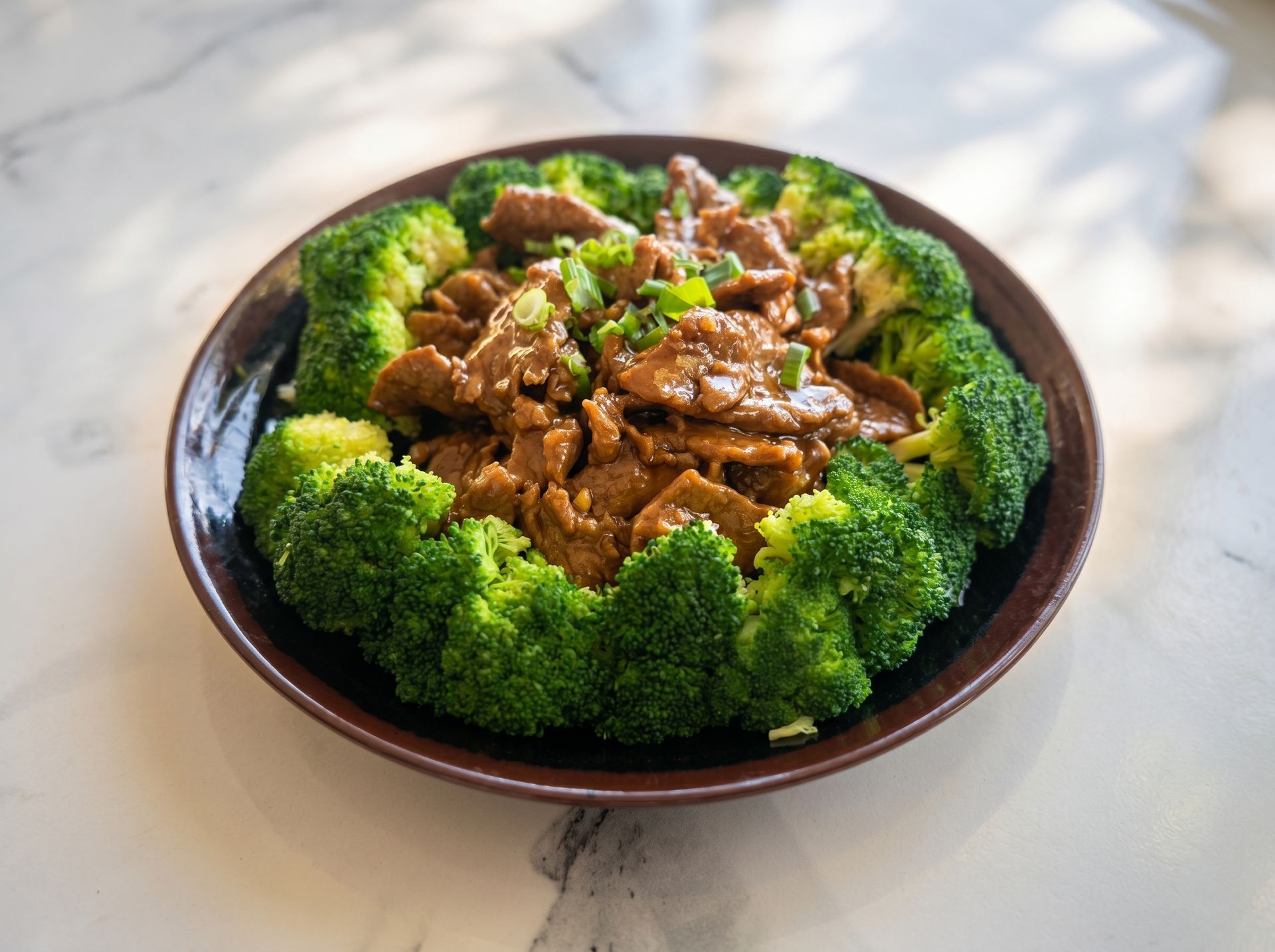Bowl of glazed meat topped with sesame seeds, surrounded by broccoli florets on a table.