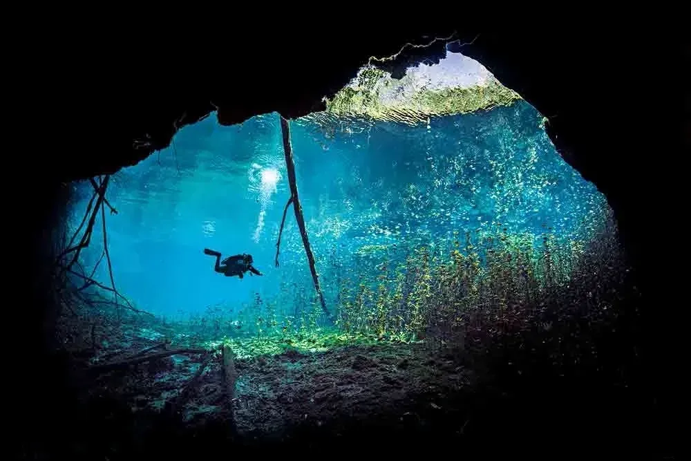 Diver swimming beneath a bright blue cave opening with sunlight filtering through the water