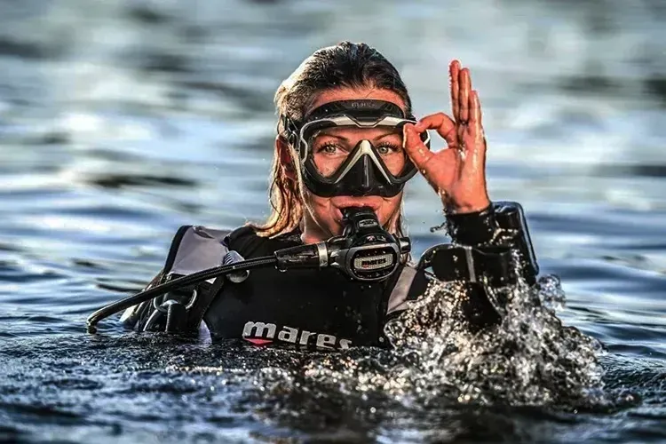 Snorkeler in black wetsuit surfacing in water, wearing mask and raising an OK hand signal.