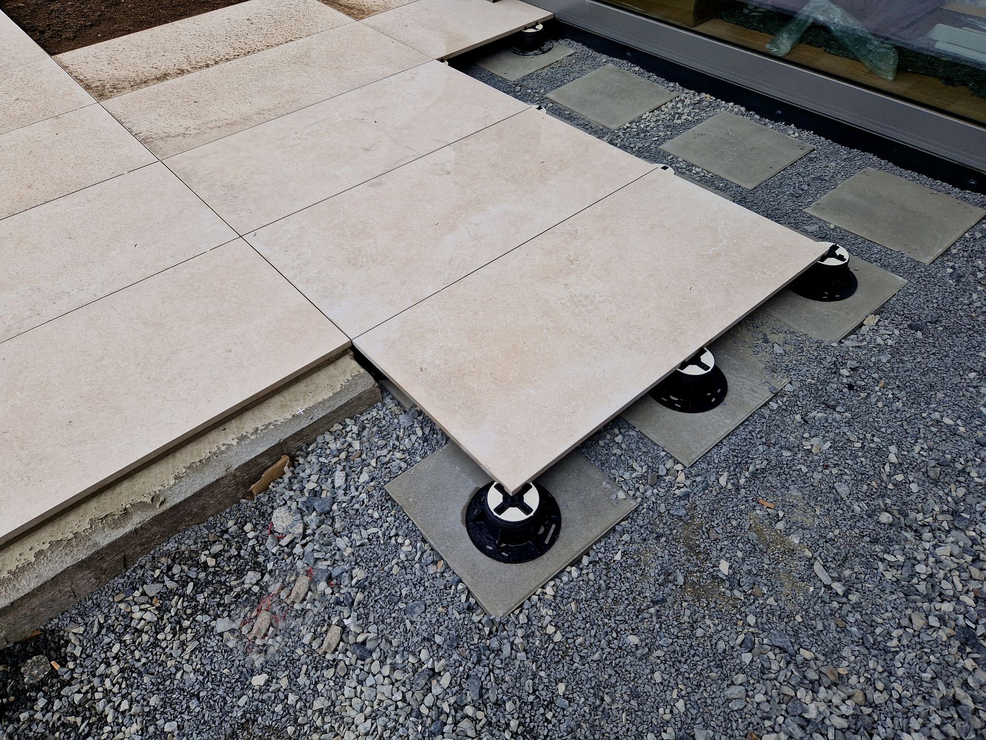 Beige tile patio partially installed with adjustable pedestals on gravel.