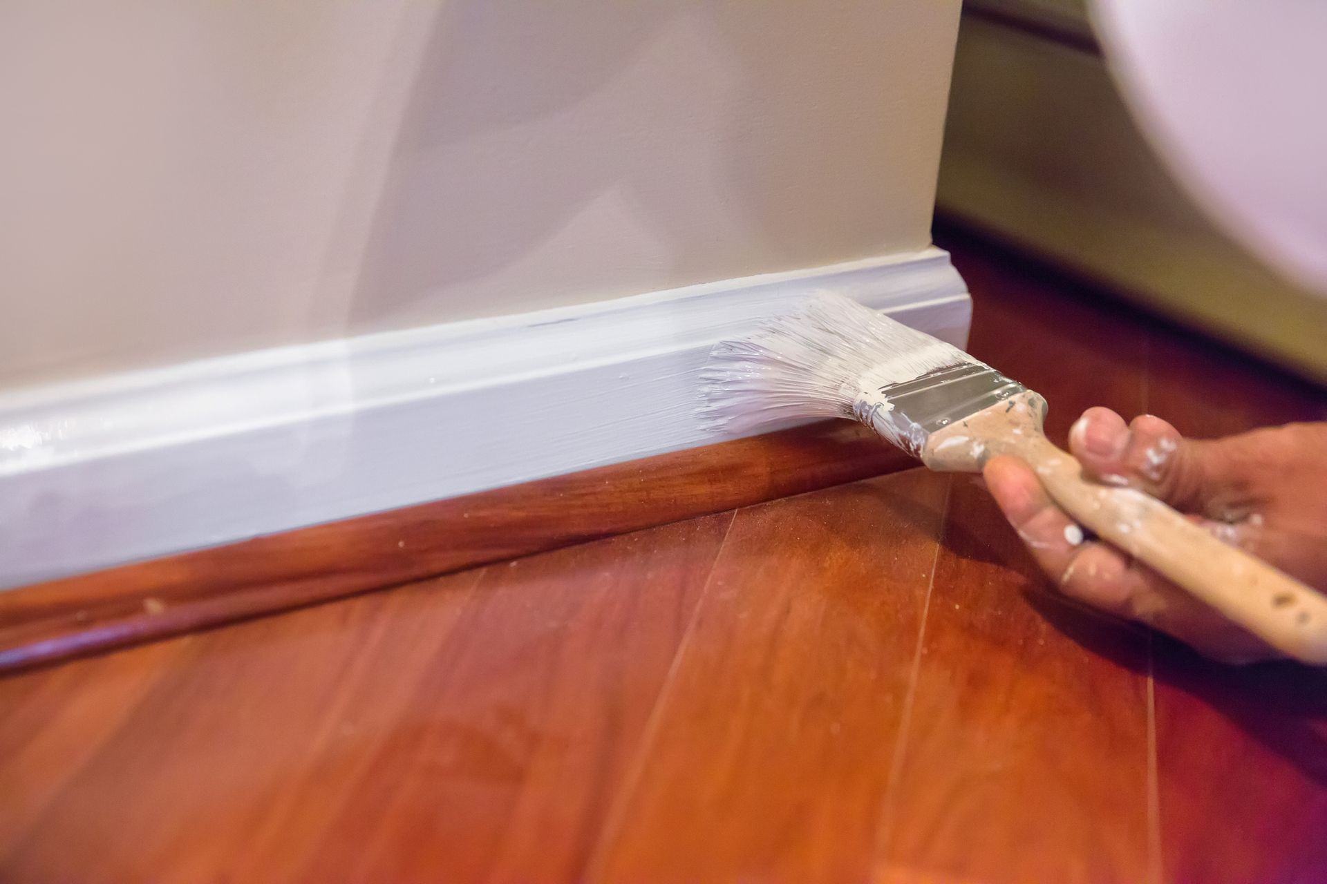 A person painting white trim on a wall above a hardwood floor with a brush.