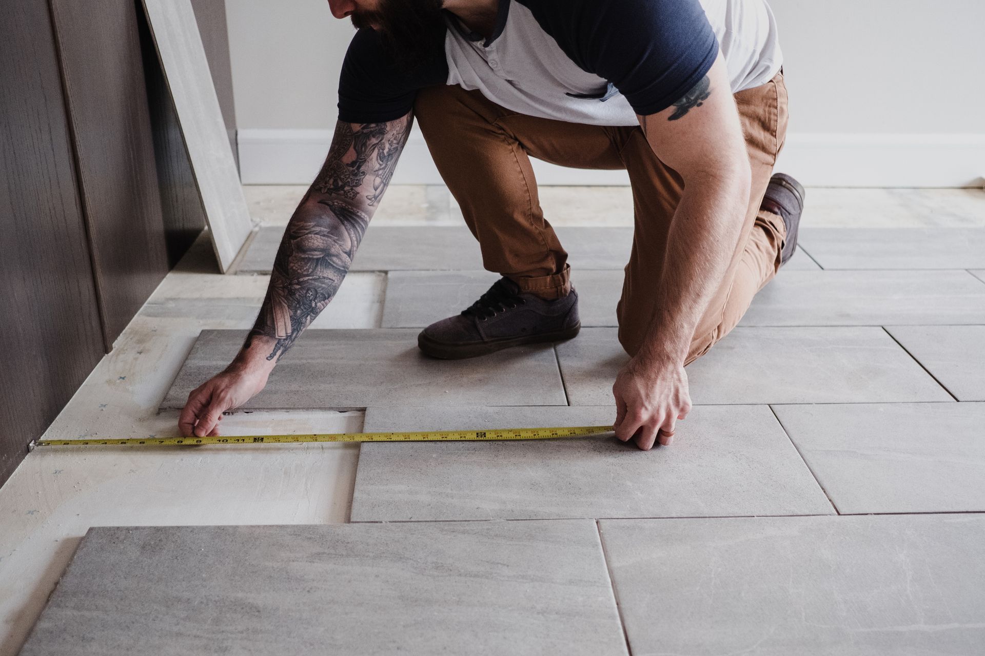 Man kneeling, measuring floor tile with a tape measure during installation.