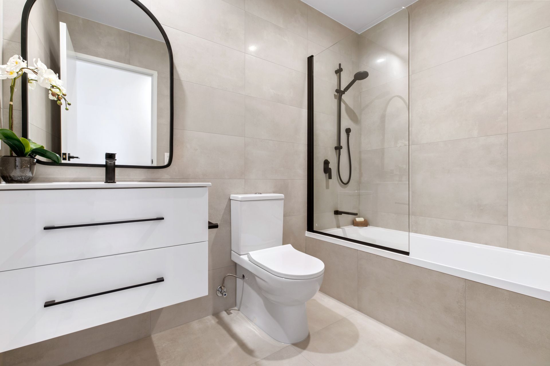 Modern bathroom with white vanity, toilet, and bathtub. Black shower fixtures, mirror, and handles. Beige tile walls and floor.