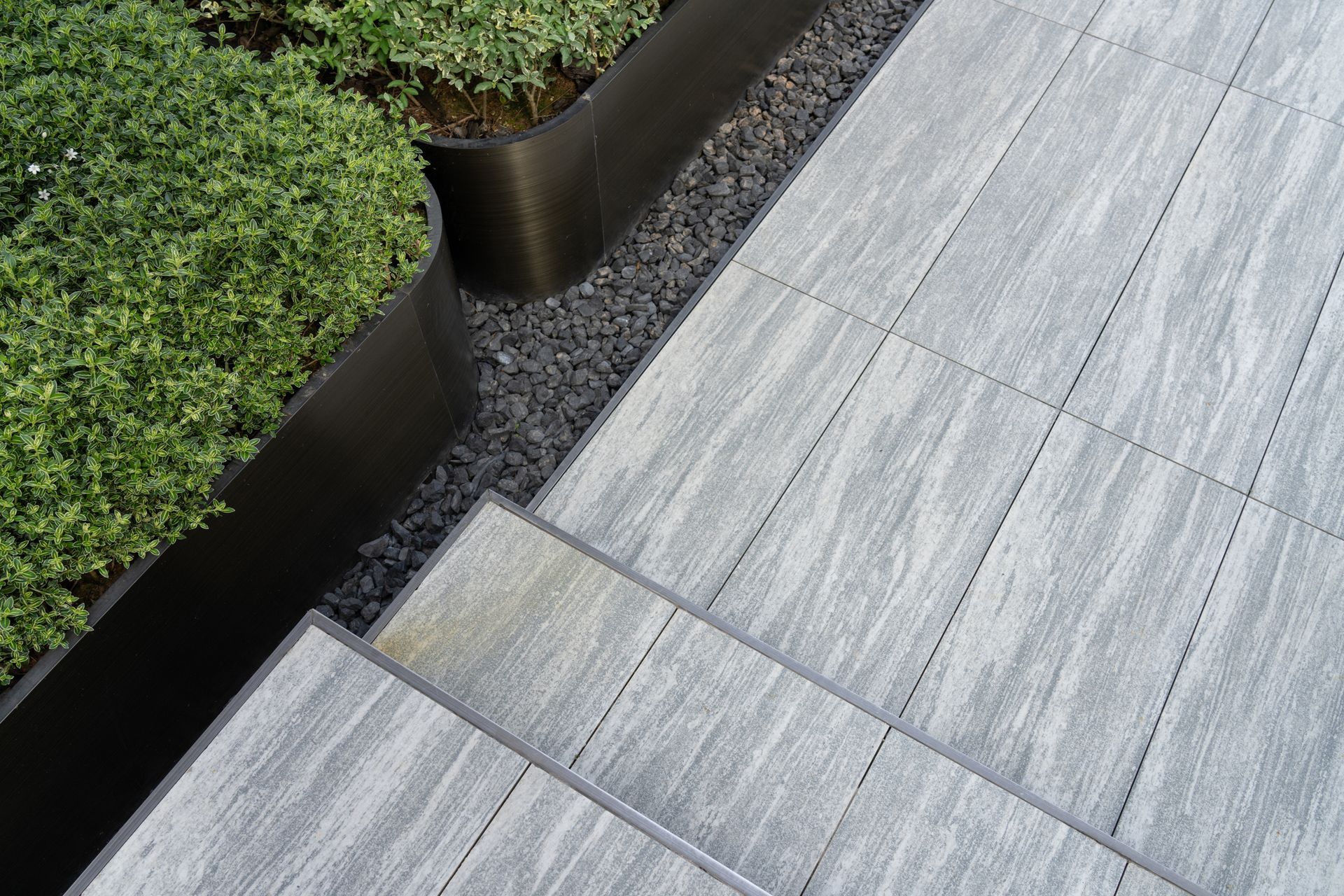 Elevated patio with light gray textured pavers and steps, next to a garden bed with dark pebbles and green plants.