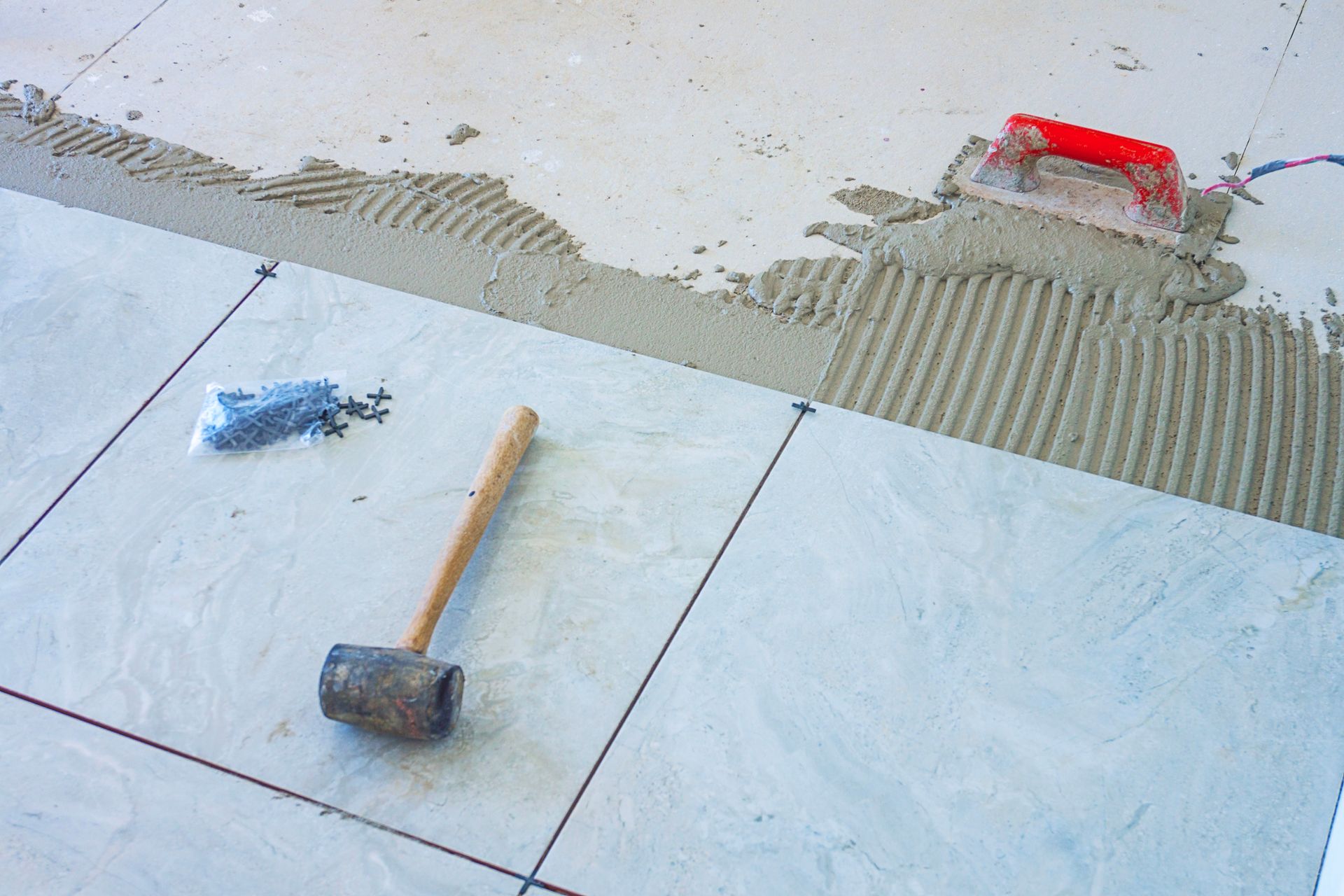 Tiling in progress: gray tiles, mortar, trowel, rubber mallet, tile spacers, on light-colored surface.