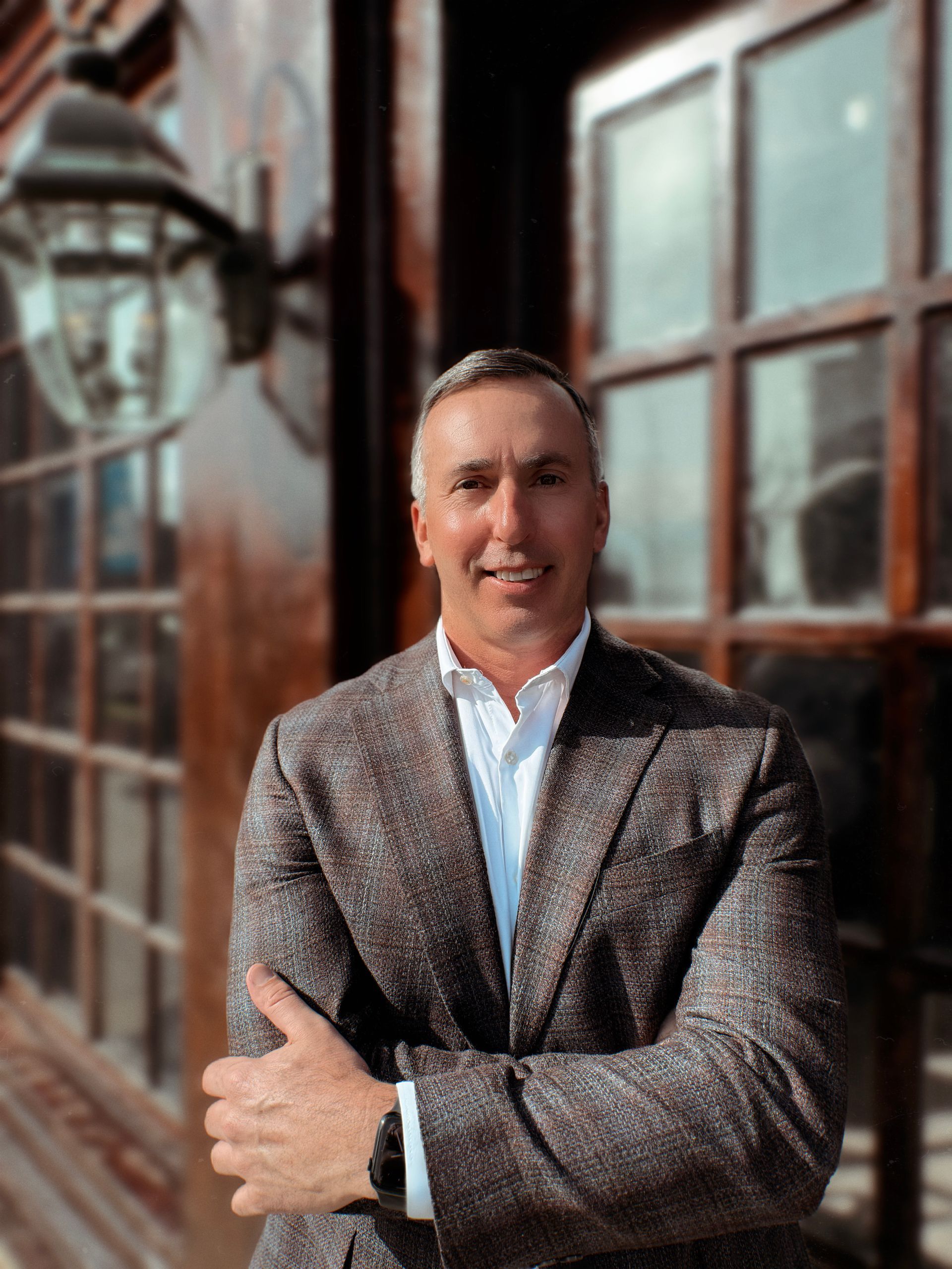 Man in patterned blazer, arms crossed, smiling, standing outside a building with windows.