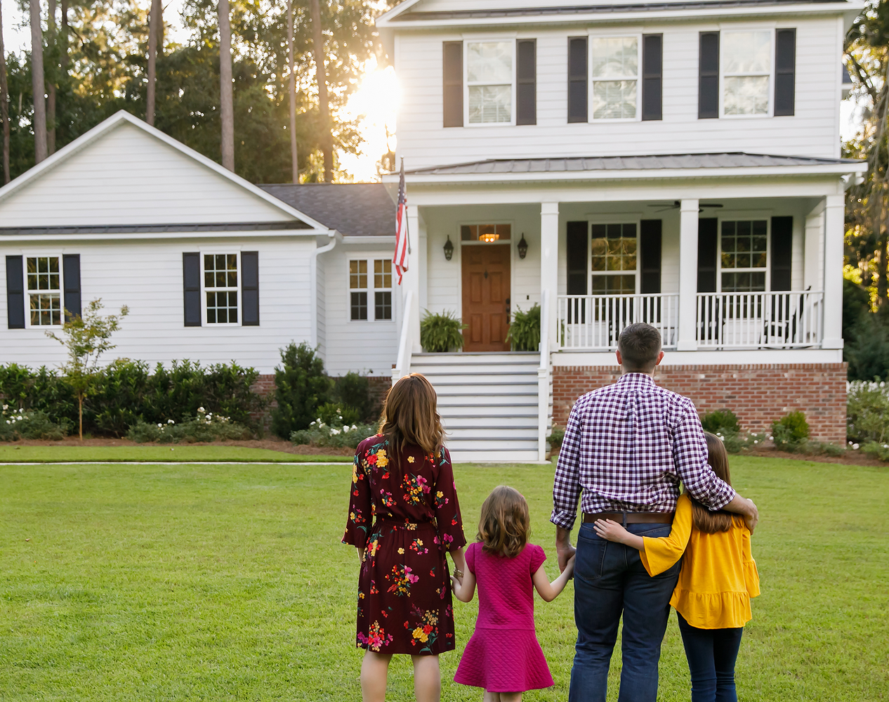 Family standing in front of a white house with green lawn, trees, and sunlight.