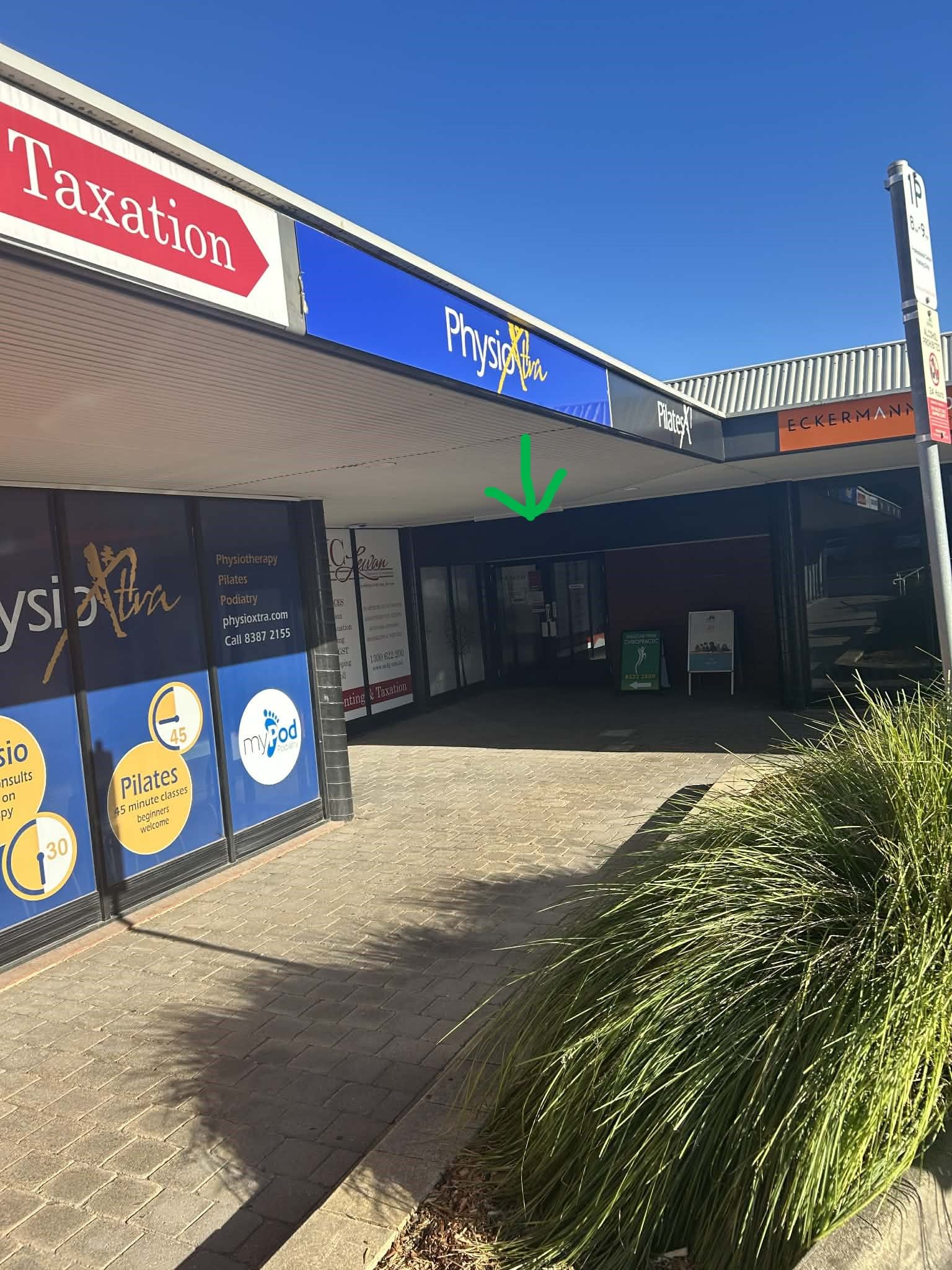 Exterior view of a shopping center with storefronts, parking, and a blue sky. A green arrow points down.