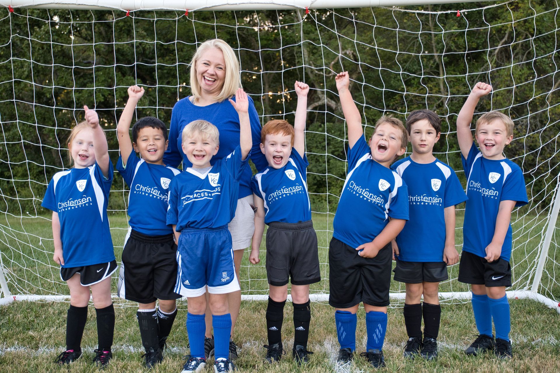 A soccer team and their coach in blue uniforms pose with their arms raised in celebration in front of a goal net.