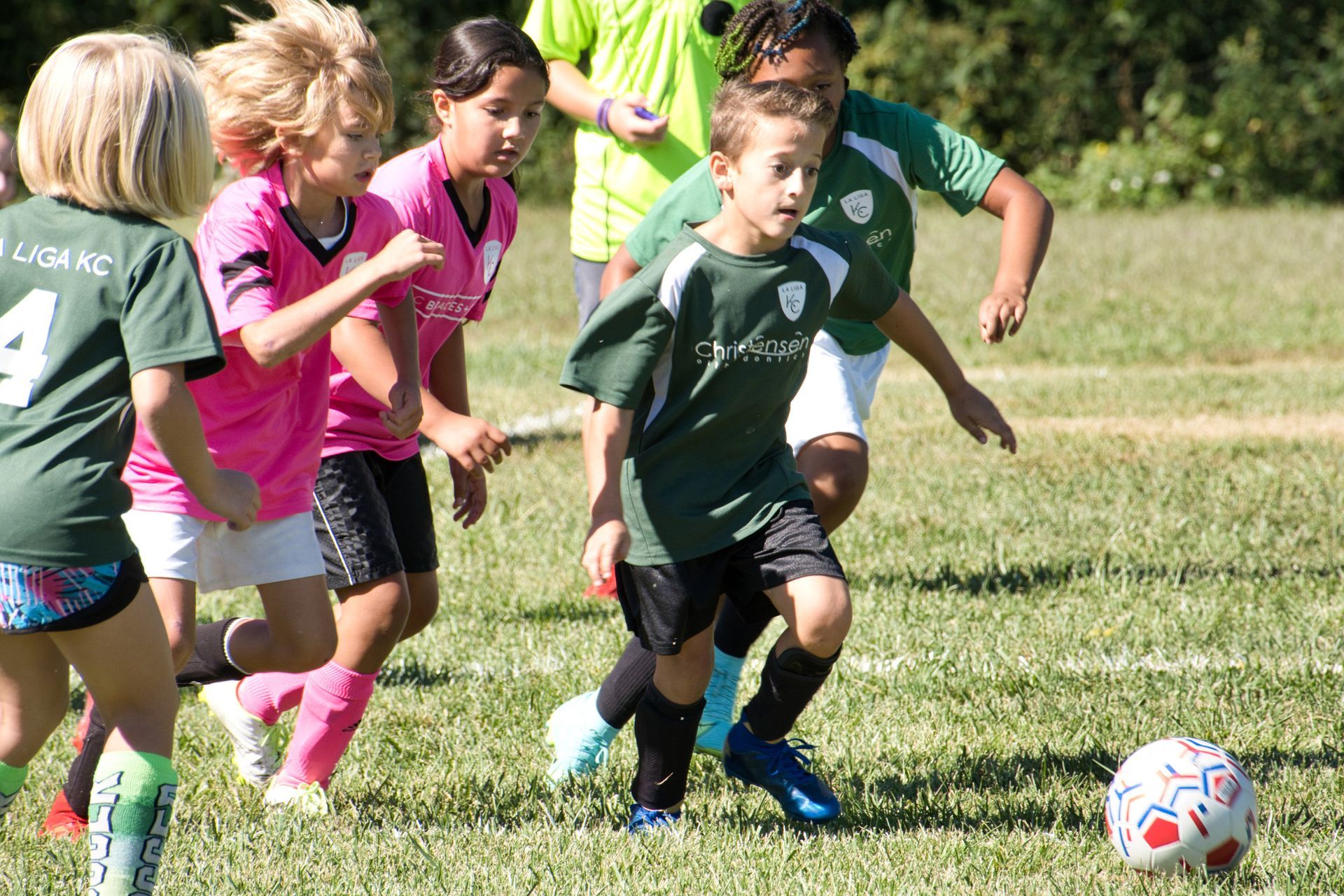 Youth soccer players in green and pink jerseys compete for a ball on a sunny grass field.
