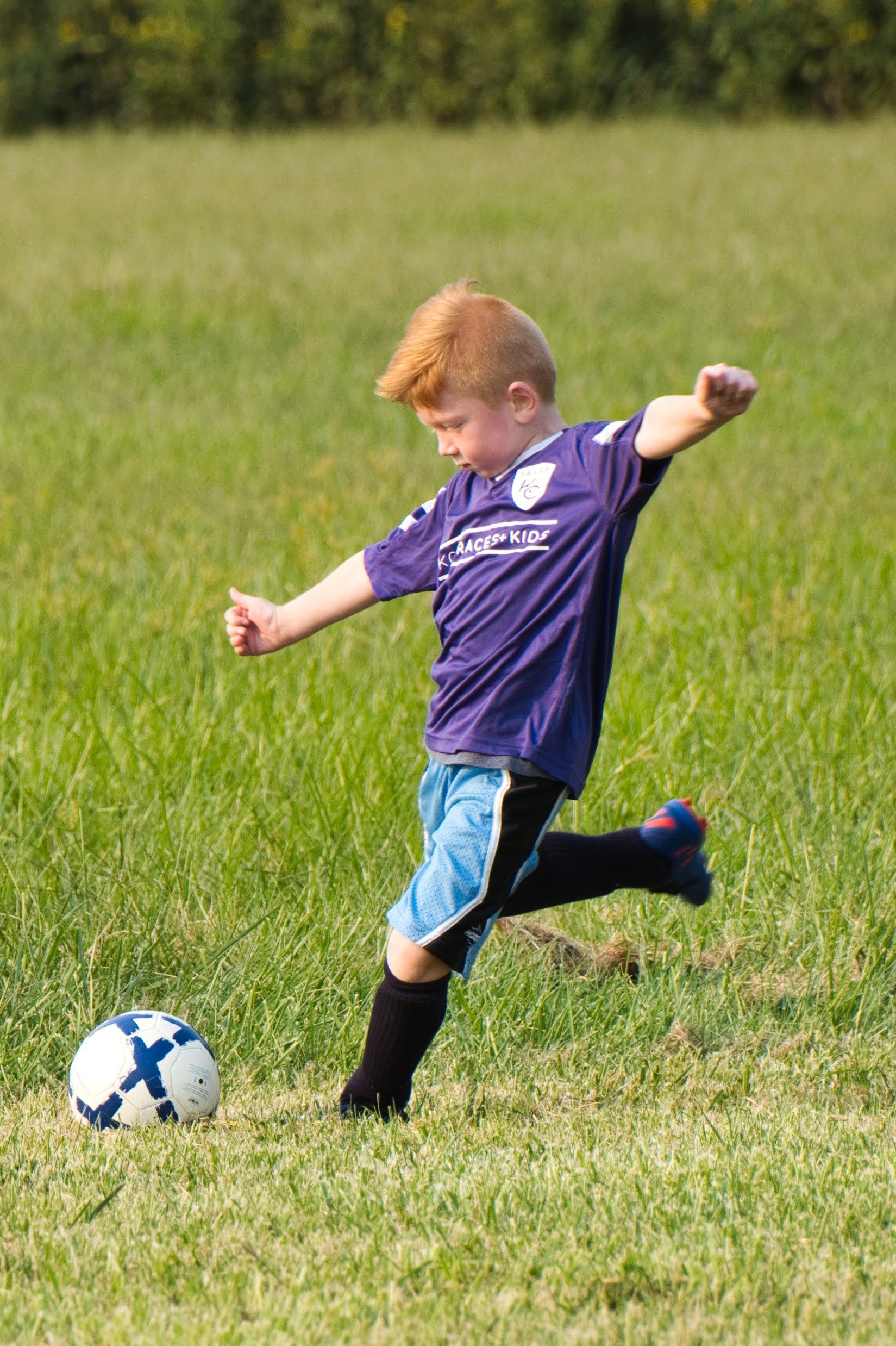 A person in a purple jersey and blue shorts kicks a soccer ball across a grassy field.