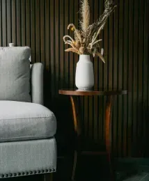 Gray armchair next to a small wooden table with a white vase holding dried flowers. Dark wooden wall behind.