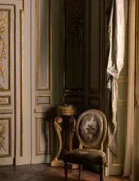 Ornate room with gold detailing, light-colored walls, patterned chair, and large window with draped curtains.
