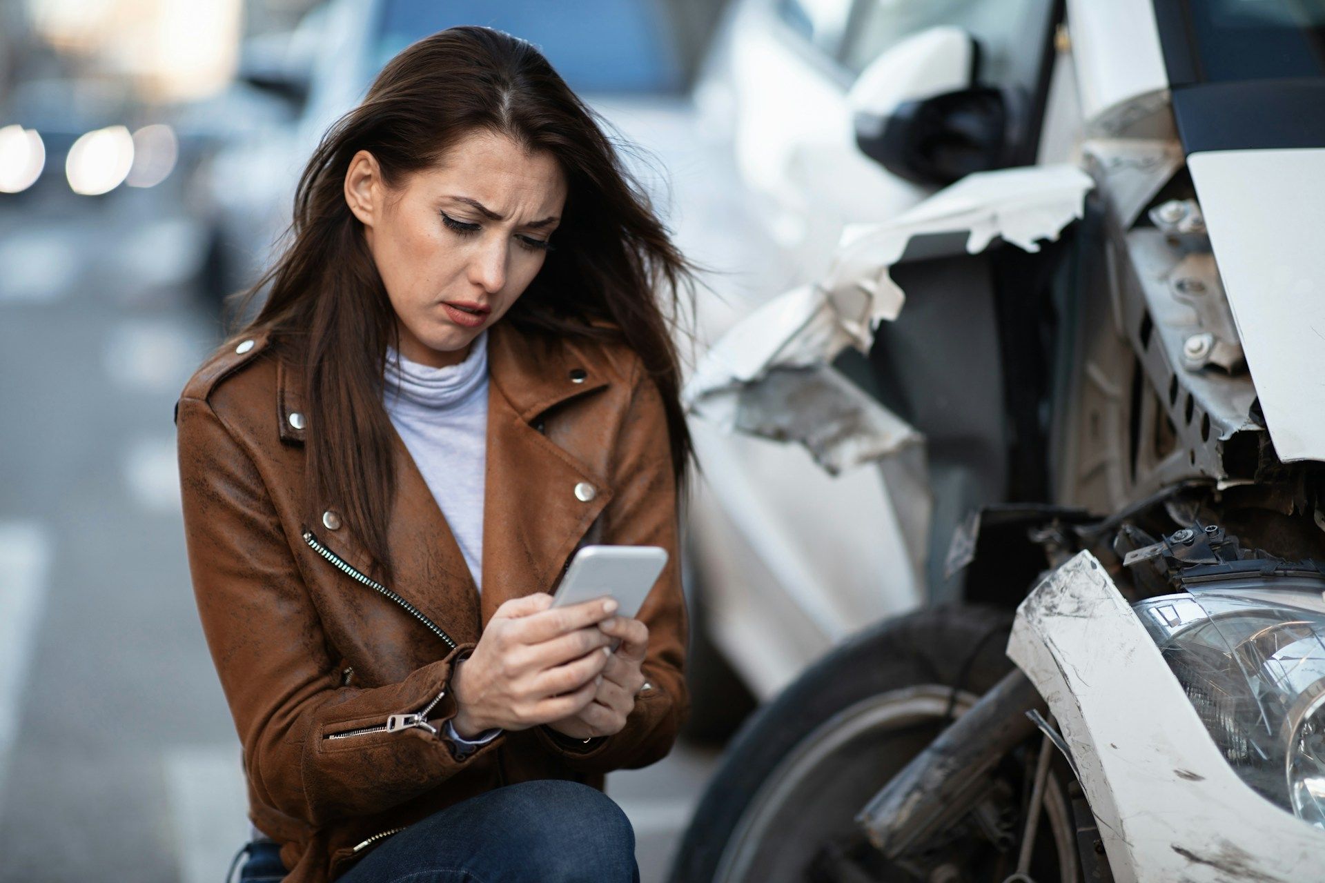 Woman in brown jacket crouches by a damaged car, looking at her phone at an accident scene
