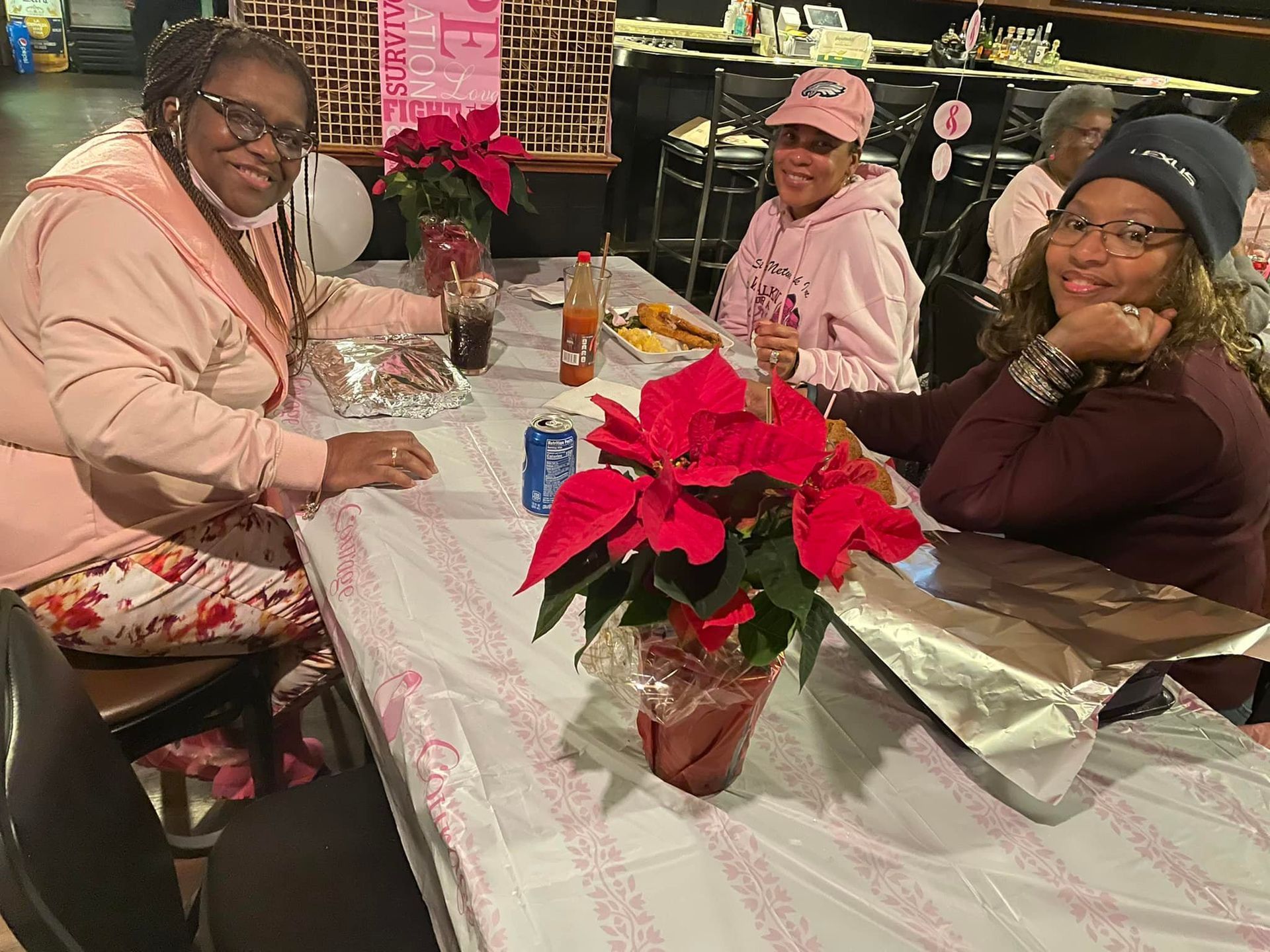 Three women in pink clothing seated at a table with food and poinsettias, likely a breast cancer awareness event.