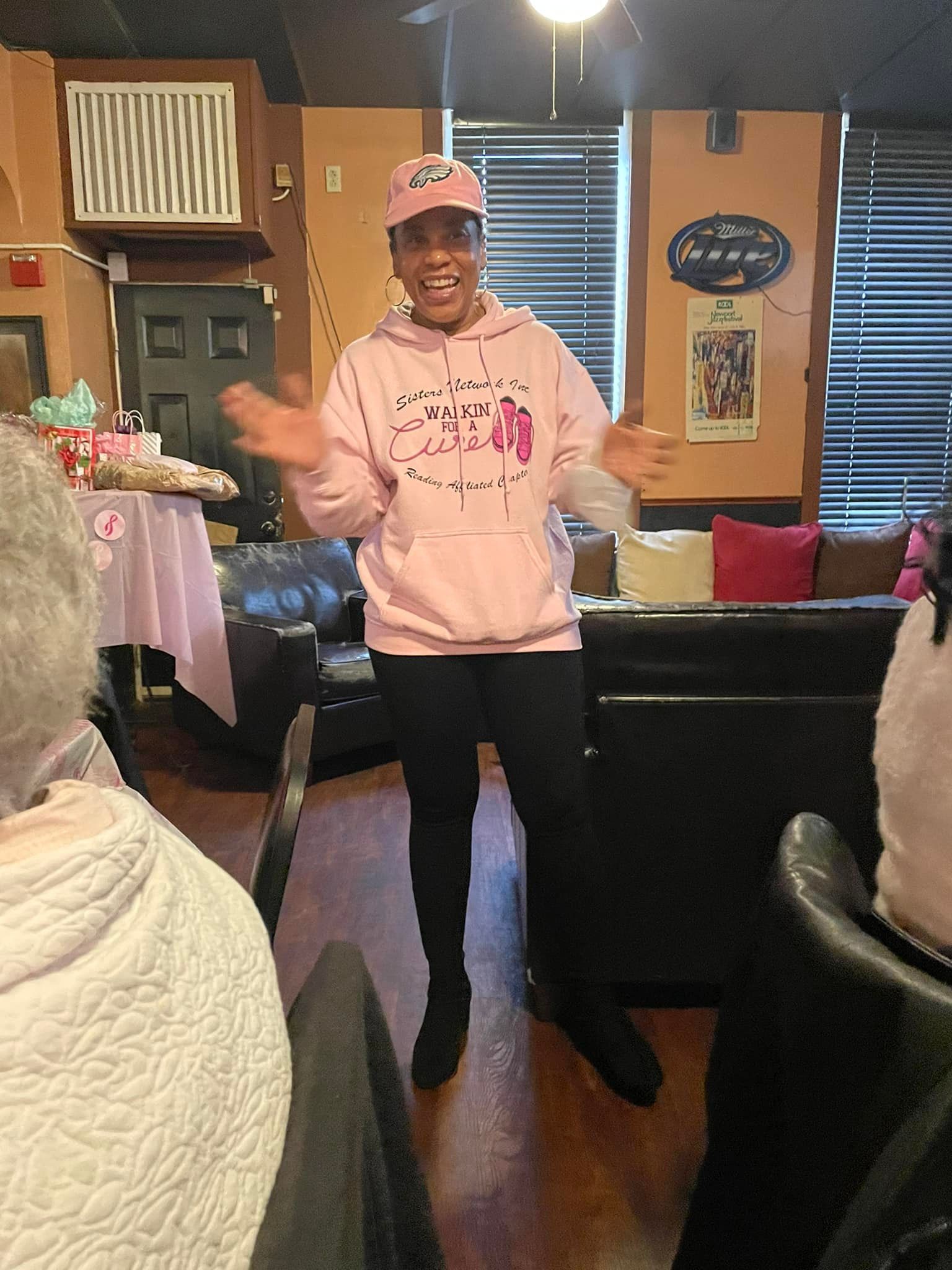 Woman in pink hoodie and cap gestures in a room with a pink-themed table.