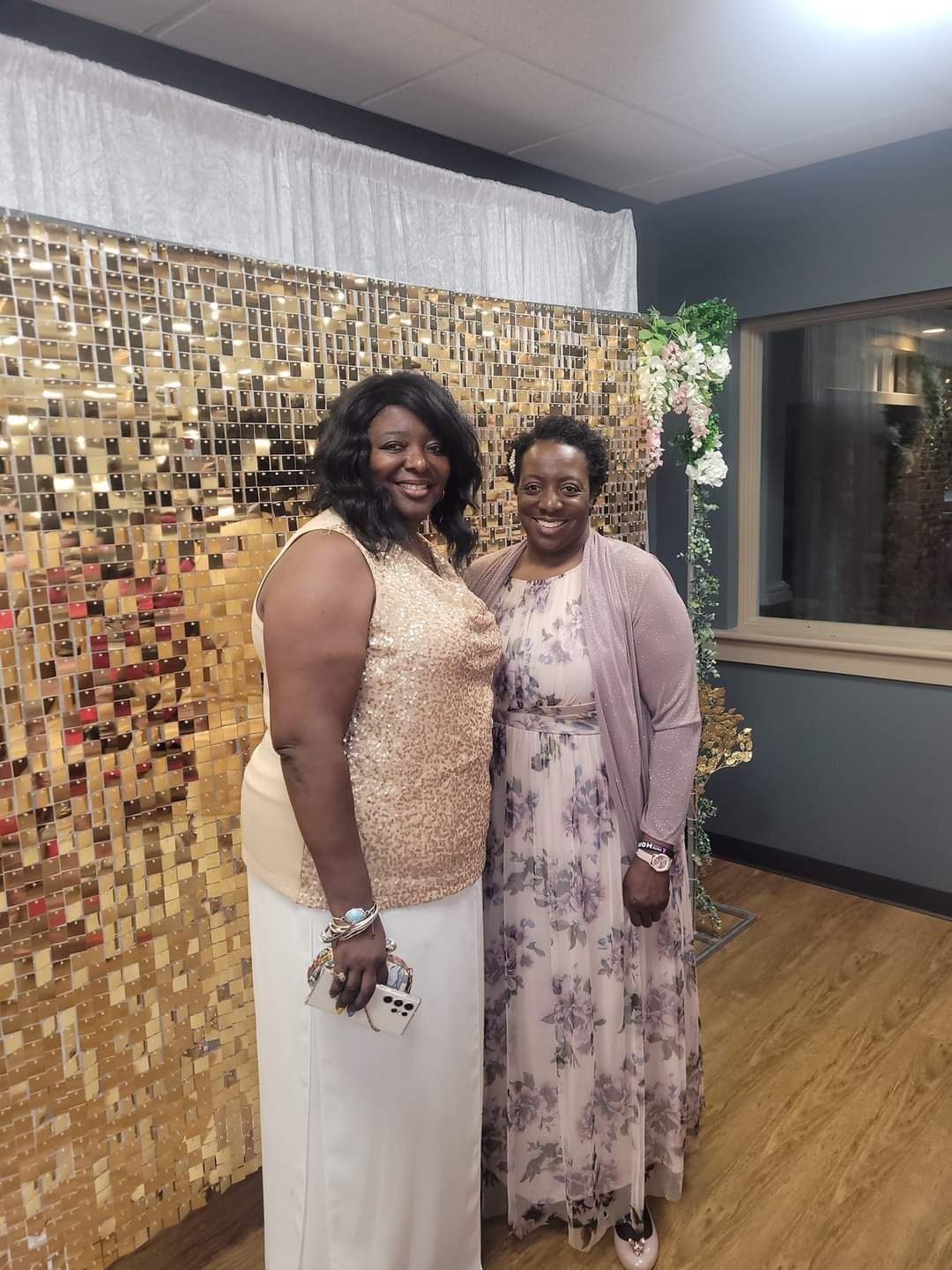 Two women smiling, posing in front of a gold sequin backdrop; one wears a gold sequined top, the other a floral dress.