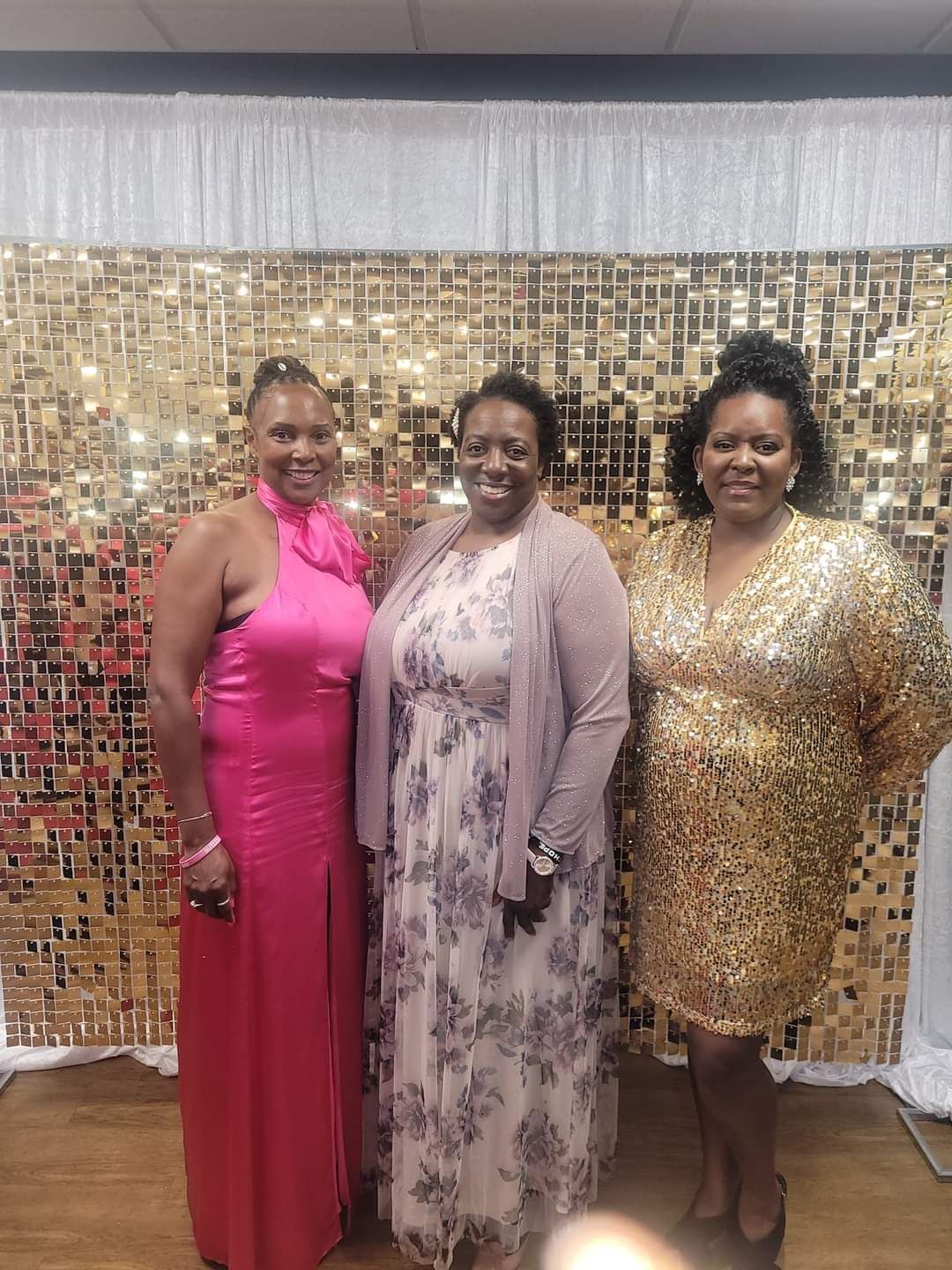Three women pose in front of a gold sequin backdrop. They are dressed in formal wear, smiling.