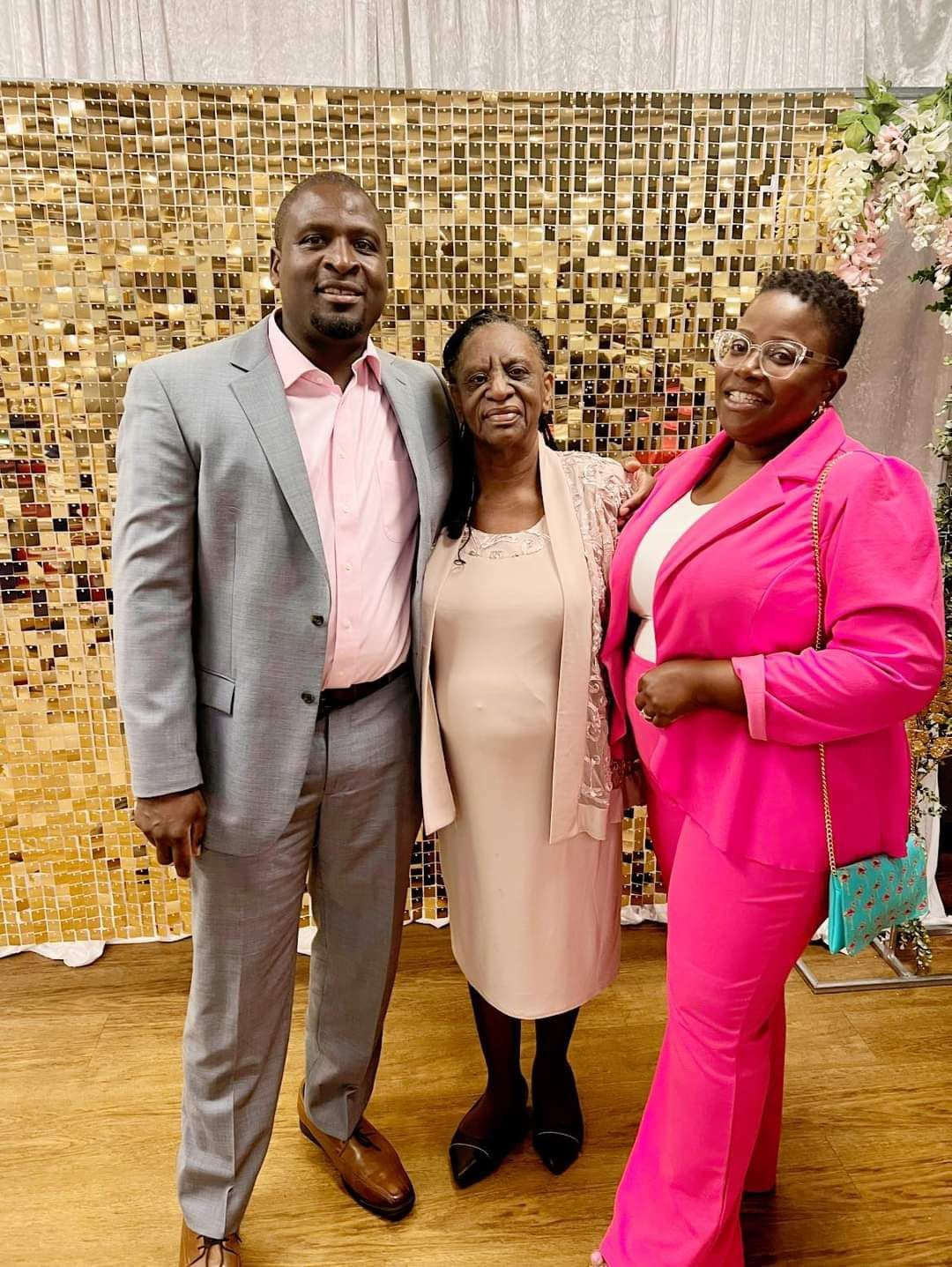 Three people posing together; man in gray suit, woman in pink dress, woman in pink pantsuit, golden backdrop.