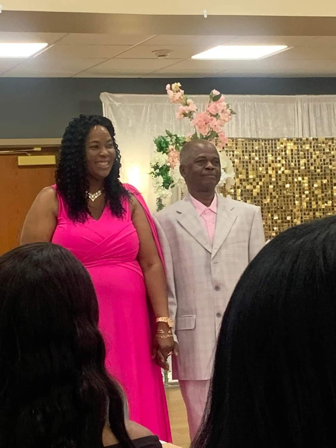 Woman in pink dress and man in light suit, holding hands at an event. Background has flowers and gold backdrop.