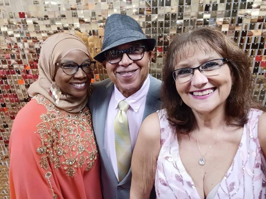 Three smiling people posing together, one in a hijab, another in a suit and hat, the third in a floral dress.