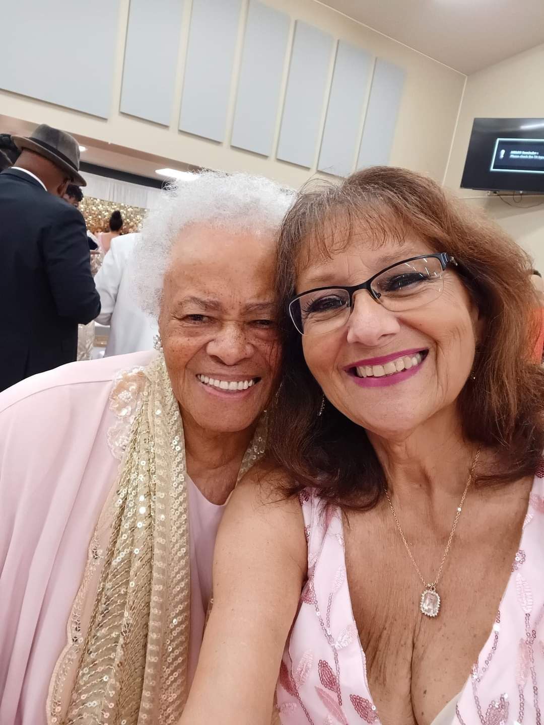 Two women smiling for a selfie. One with white hair, the other wearing glasses. Both in pink.