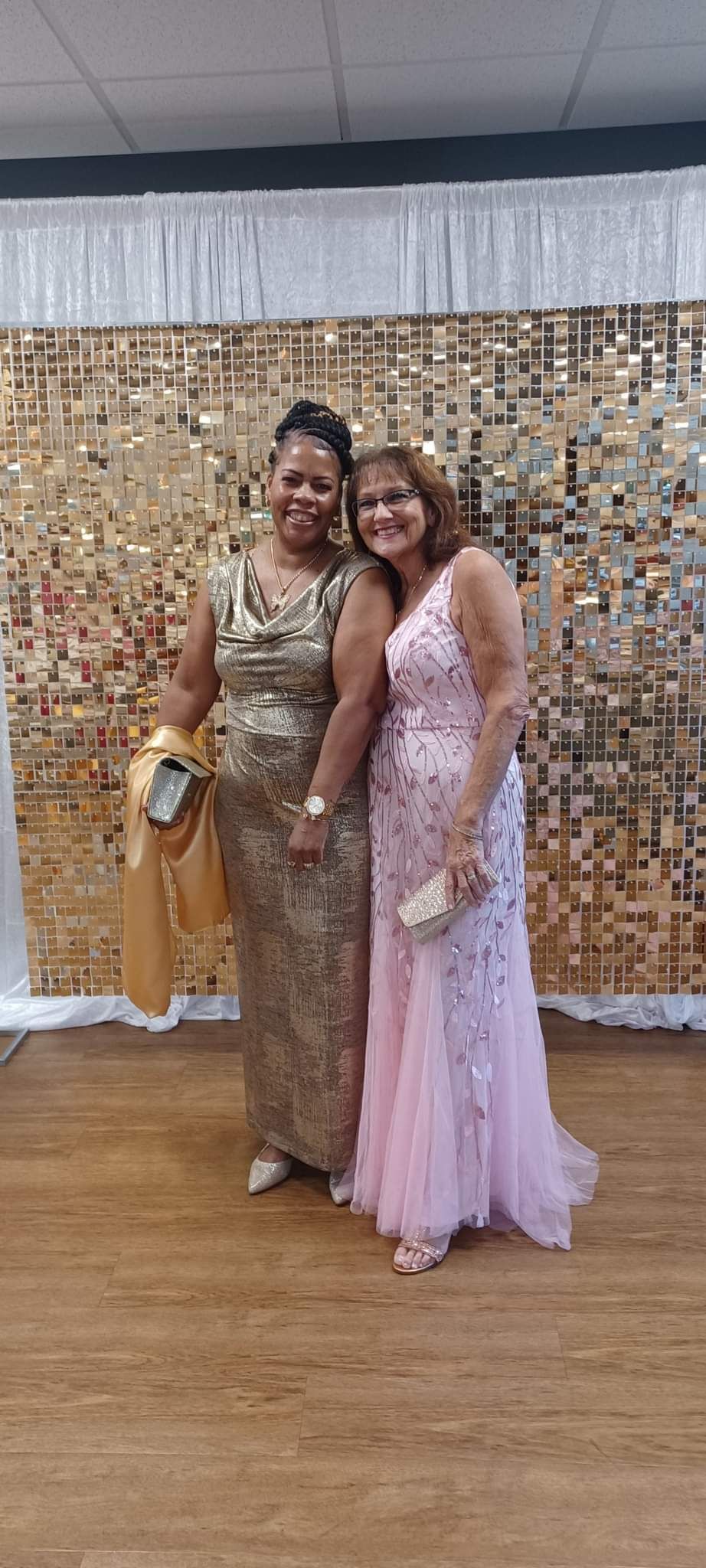 Two women in evening gowns pose in front of a sequin backdrop. One in gold and another in pink.