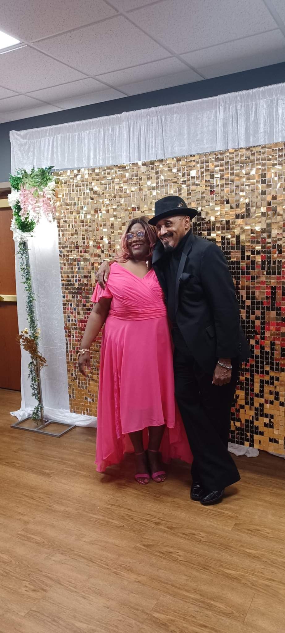 A smiling couple poses in front of a gold sequin backdrop. The woman wears a pink dress. The man wears a black suit and hat.